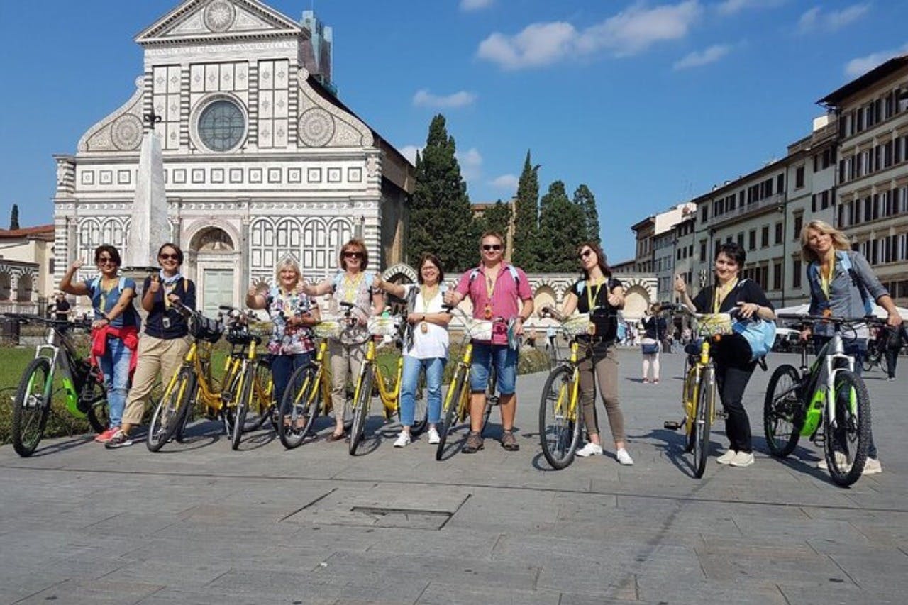 A group of people with yellow bicycles stands in front of an ornate building with trees in the background on a sunny day.
