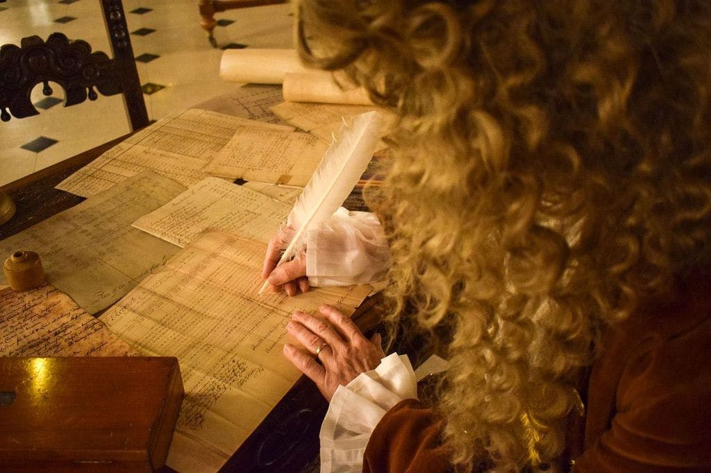 一个头发卷曲的人在木桌上用羽毛笔写字，周围是卷轴和文件。