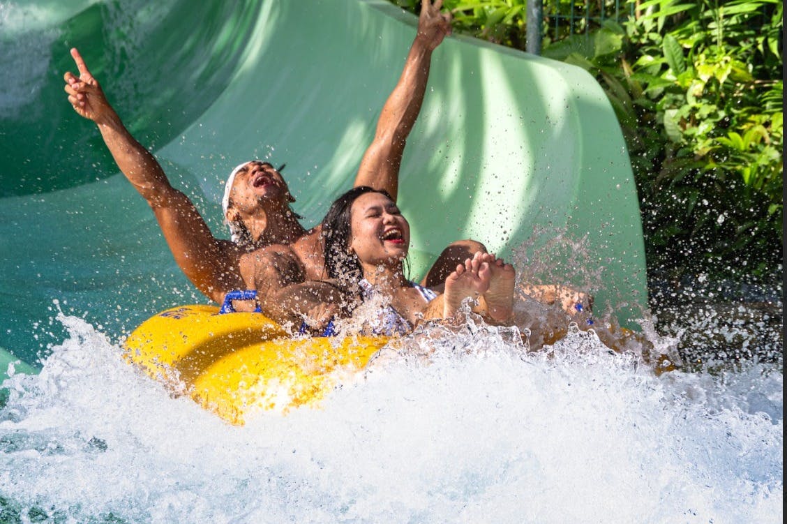 Twee mensen rijden op een geel opblaasbaar vlot van een waterglijbaan, lachen en spetteren water.