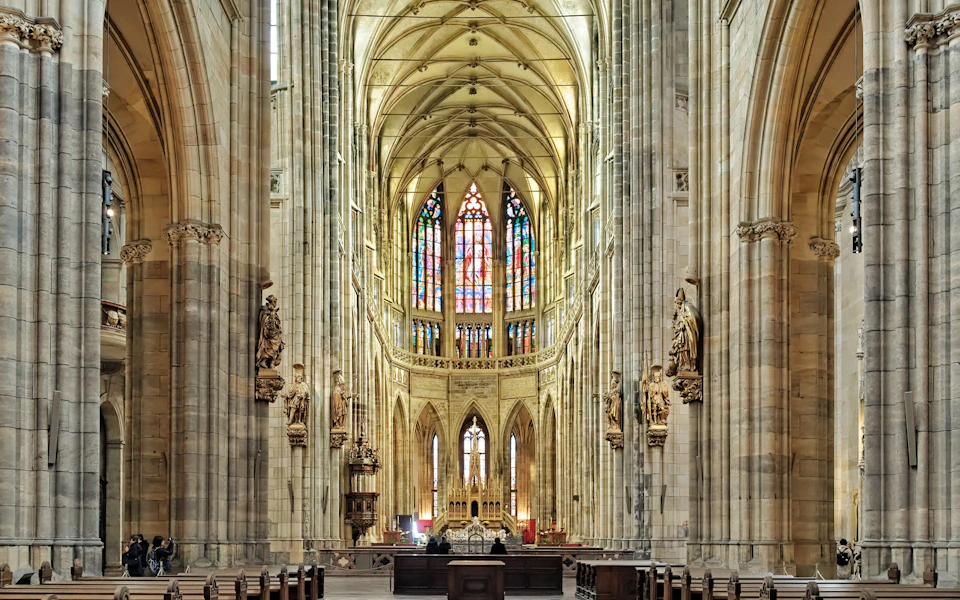 Interior of a grand cathedral with vaulted ceilings, statues, wooden pews, and a large stained-glass window at the front.