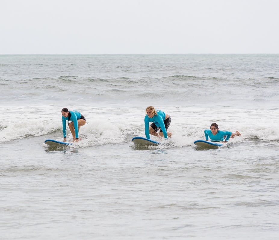 Tři lidé v modrých neoprenech surfují v oceánu na malých vlnách na modrých surfových prknech.