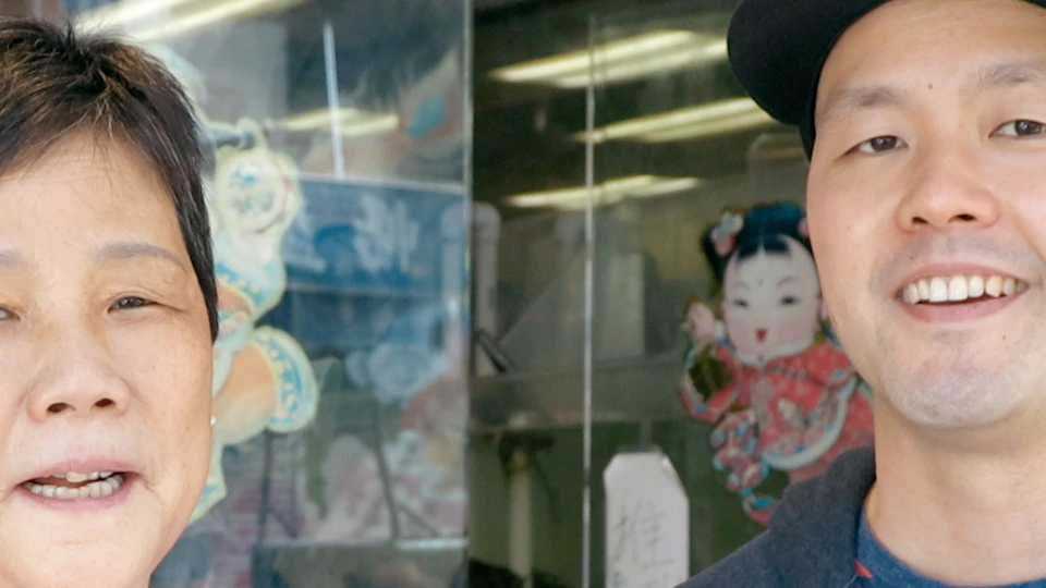 Two people standing in front of a storefront; one wears a white apron and the other a blue shirt and black cap.