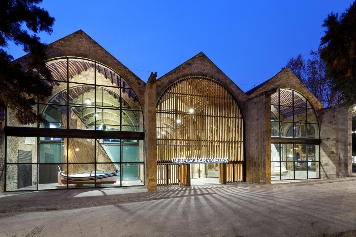 Bâtiment moderne à façade vitrée avec fenêtres cintrées, entrée en lattes de bois et exposition de bateaux à l'intérieur. L'éclairage du soir accentue la scène.
