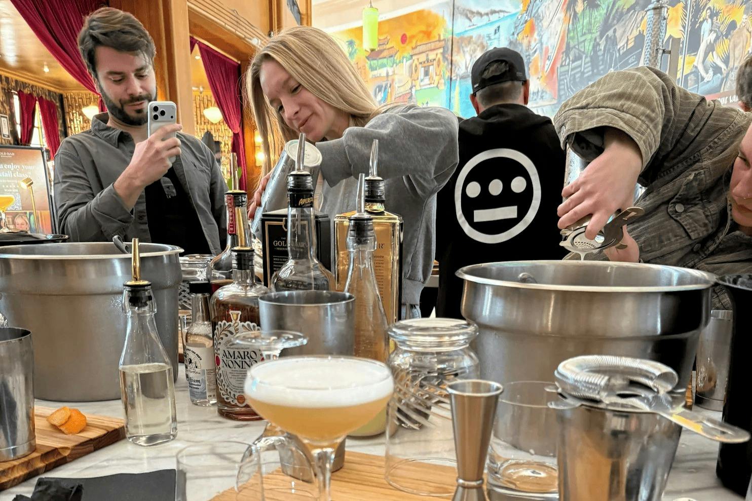 Two people at a bar station with various bottles and cocktail-making tools, one recording with a phone, the other making a drink.