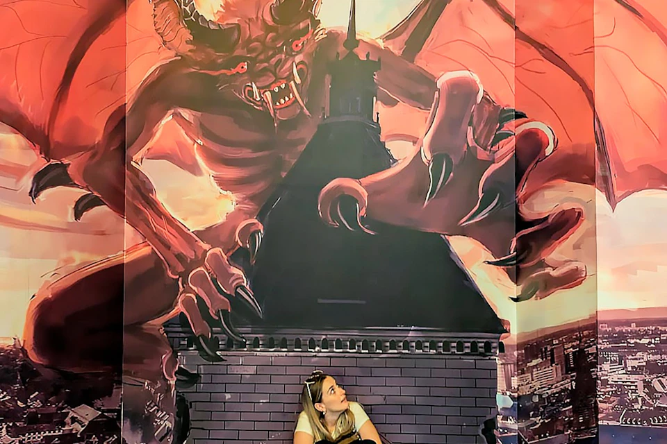 A woman sits below a mural depicting a large red demon with wings and claws looming over a building.