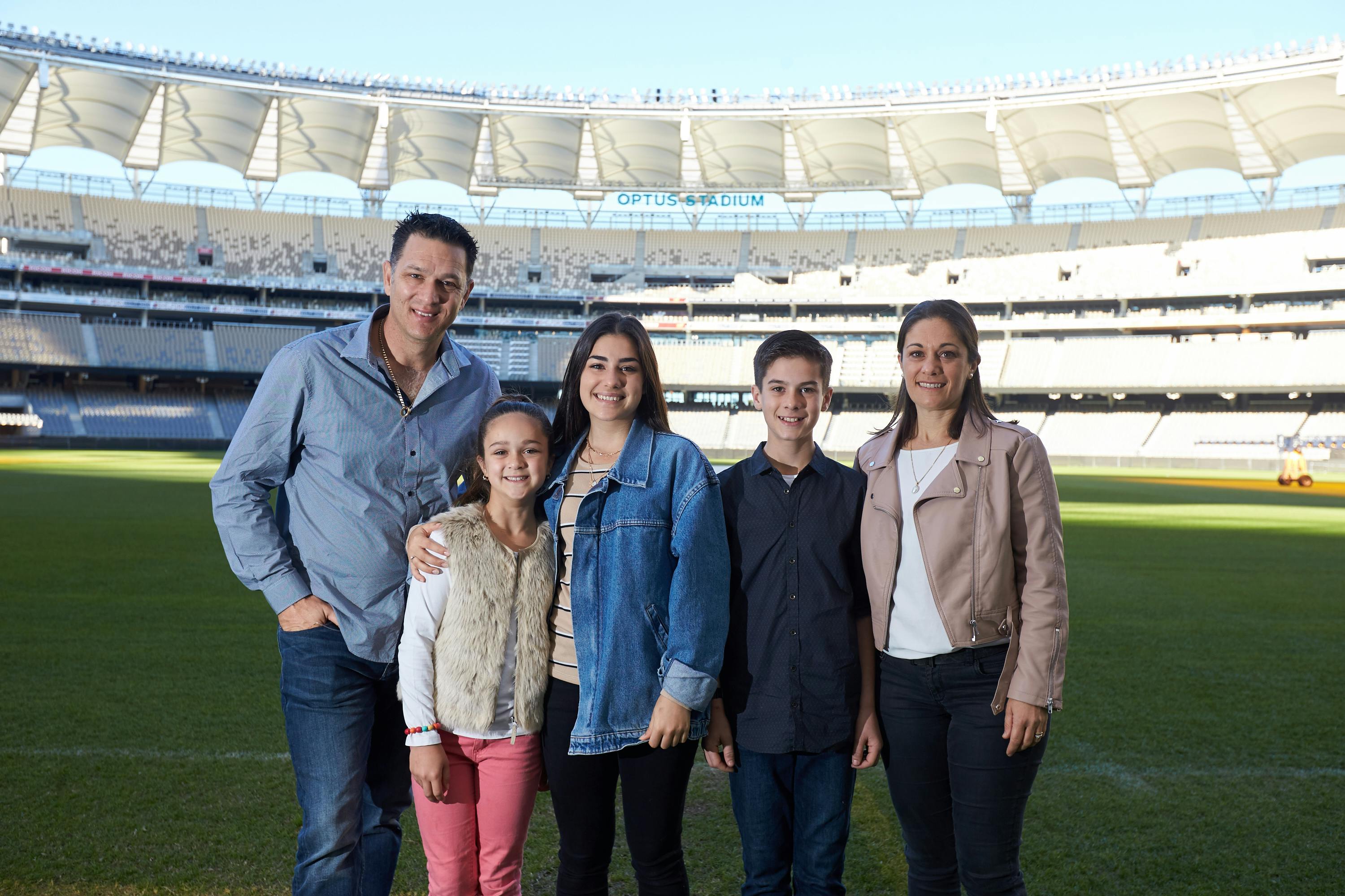 Πέντε άτομα στέκονται σε ένα χορταριασμένο χωράφι σε ένα άδειο στάδιο, χαμογελώντας στην κάμερα, με τα καθίσματα του σταδίου να φαίνονται στο παρασκήνιο.