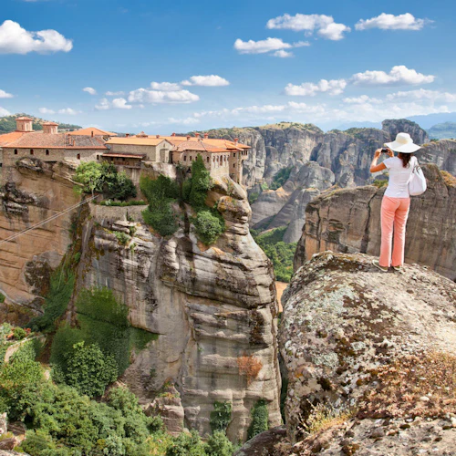 Eine Frau in rosa Hosen und weißem Hut fotografiert ein Kloster auf einer steilen Felsformation in einer Berglandschaft.