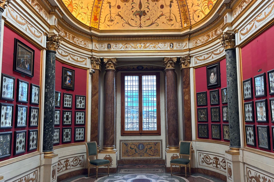 A lavishly decorated circular room with ornate walls, a domed ceiling, framed artworks, and a window offering a scenic view.