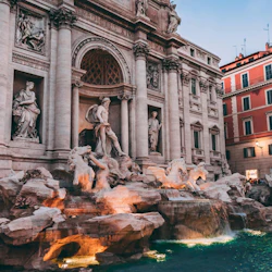 A grand Baroque fountain with statues and flowing water, set against an ornate building façade during dusk.