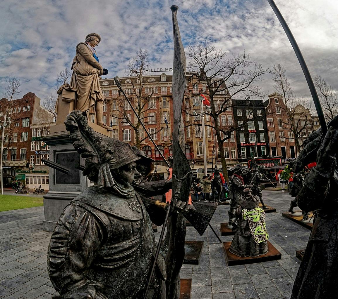 Bronze statues of armed soldiers in a plaza, with a tall statue of a man in the background and buildings behind them.