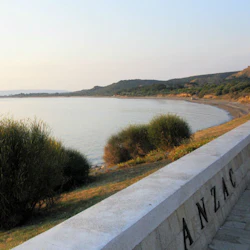 En stenmur märkt "ANZAC" har utsikt över en lugn vik med gräsbevuxna buskar och avlägsna kullar under en klar himmel.