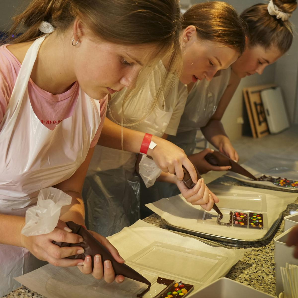 Three women decorating chocolate bars with icing and colorful candies in a kitchen setting. They are wearing aprons and focused.