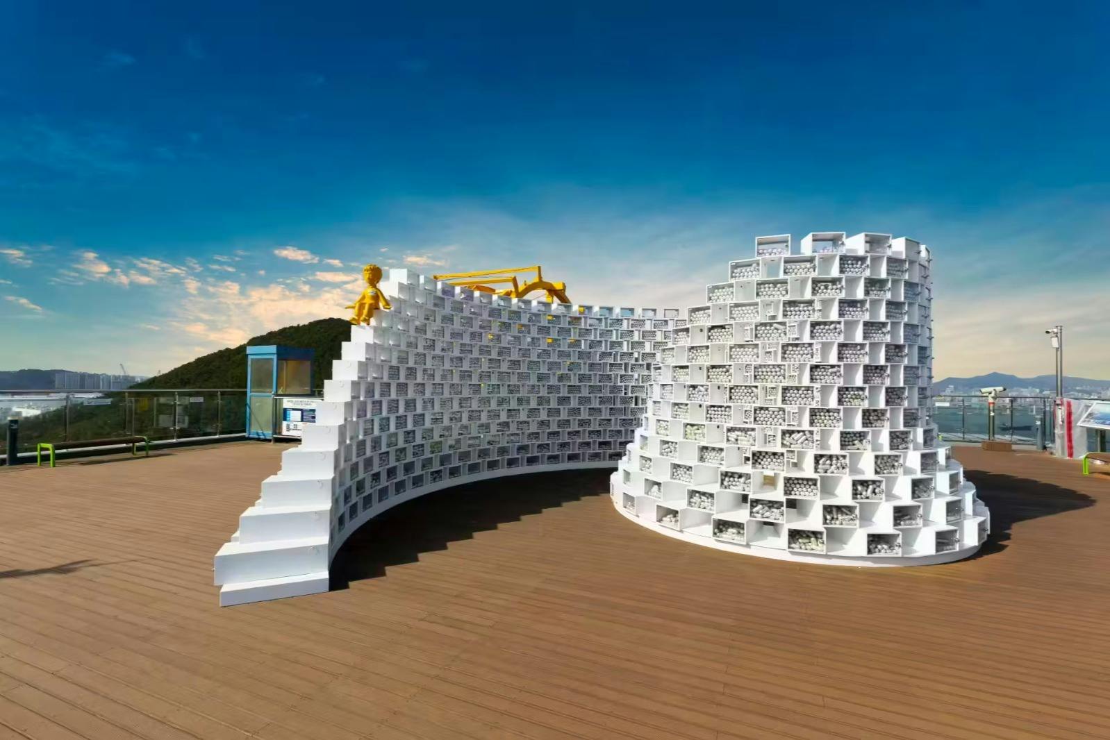 A white, curved, lattice-like structure with a staircase on a wooden deck under a blue sky, featuring yellow figures on top.