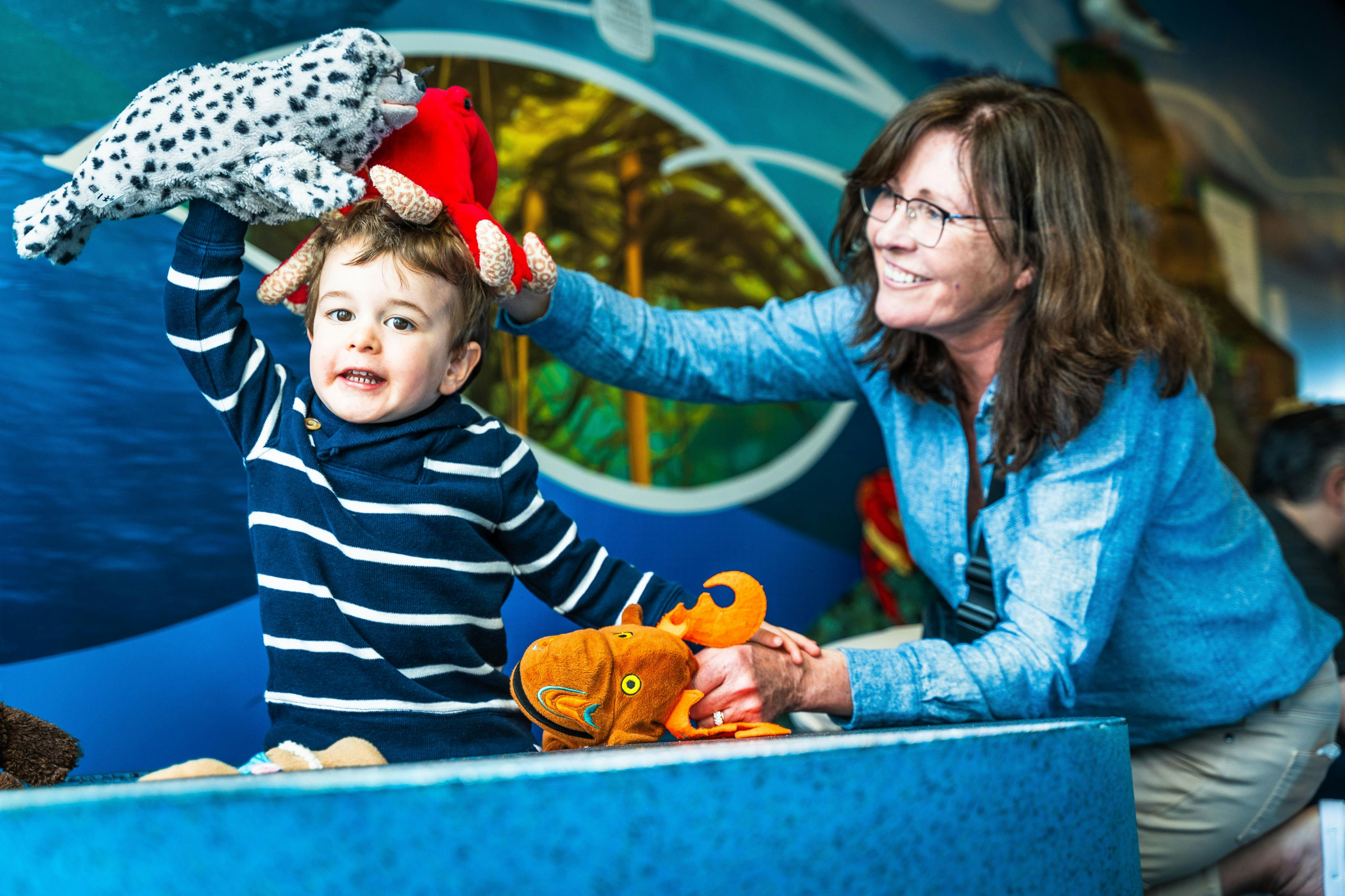 Et barn i en stribet skjorte holder et udstoppet dyr, når en voksen smiler, og justerer et andet plyslegetøj på hovedet i farverige omgivelser.