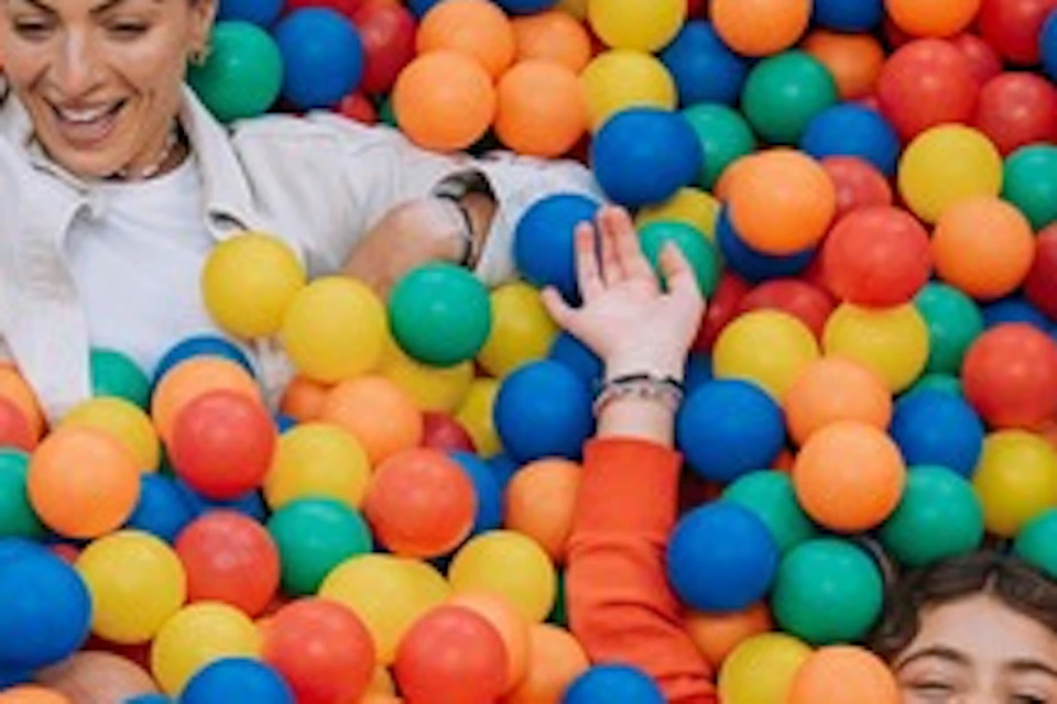 Several people smiling and playing in a large net filled with colorful plastic balls outdoors.