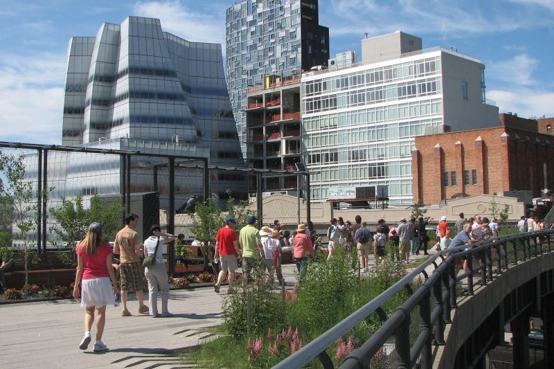 Persone che camminano su una passerella urbana sopraelevata circondata da edifici moderni e dal verde in una giornata di sole.