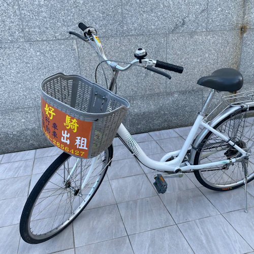 A white bicycle with a basket featuring a sign in Chinese characters, parked on a tiled surface against a stone wall.