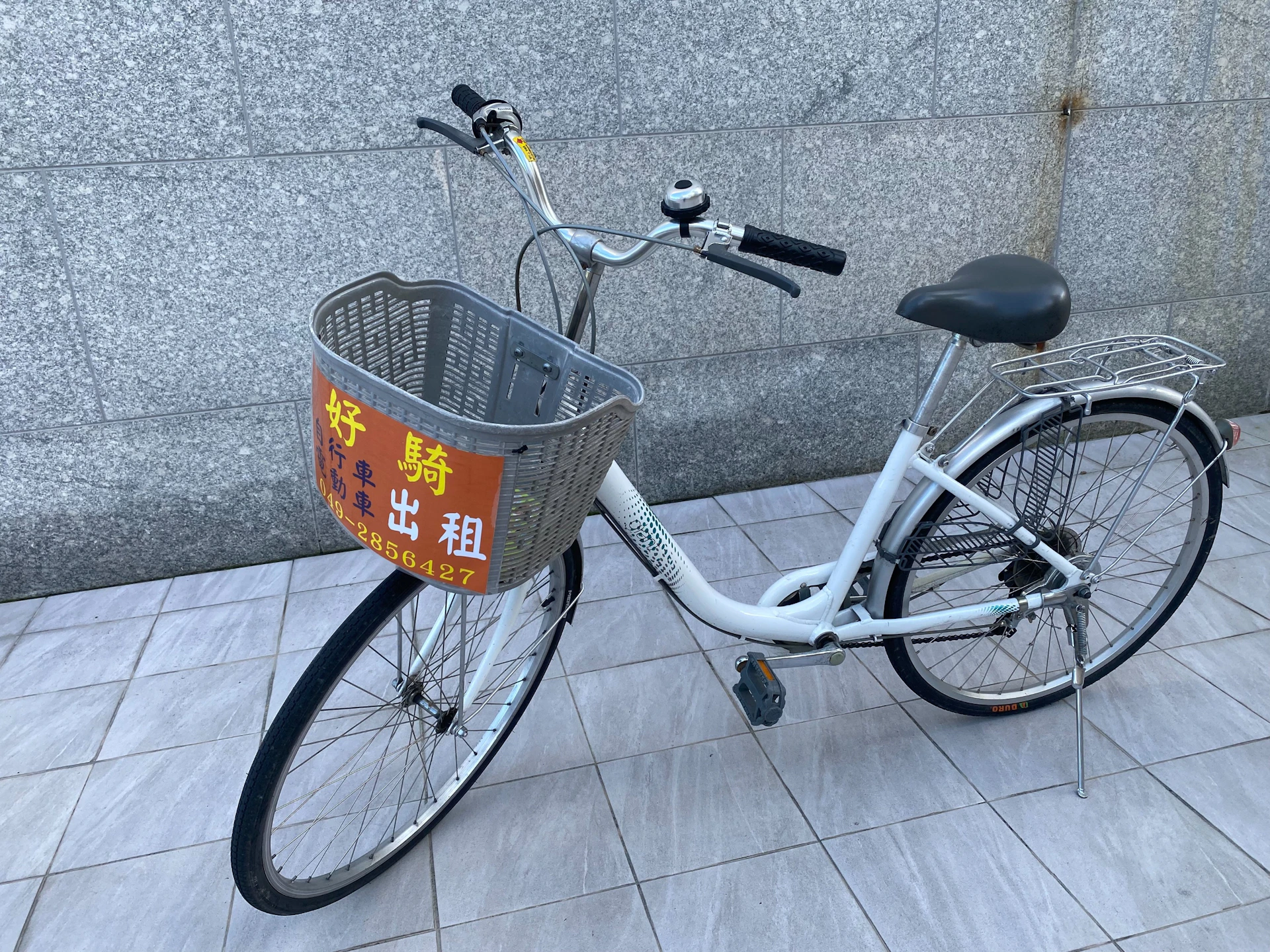 A white bicycle with a basket featuring a sign in Chinese characters, parked on a tiled surface against a stone wall.