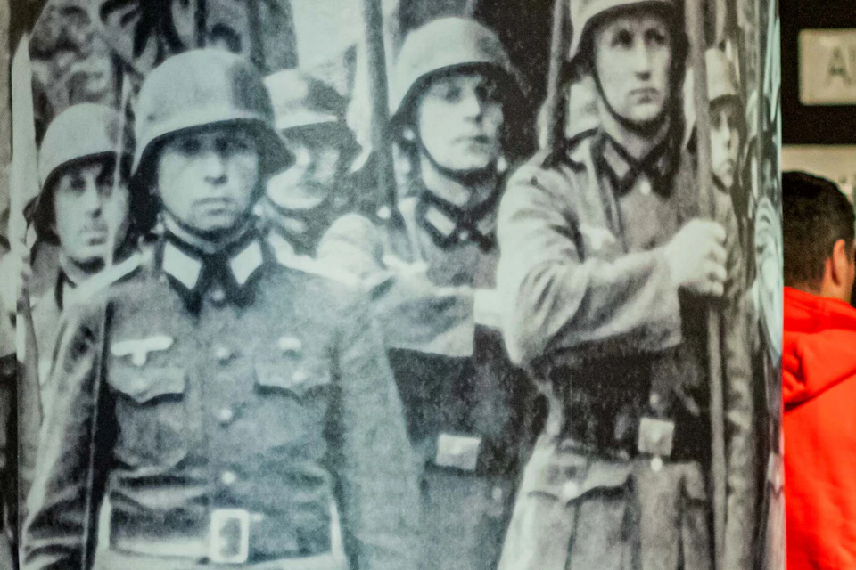 People observe and discuss exhibits in a museum, with a large black-and-white photo of soldiers marching on one wall.