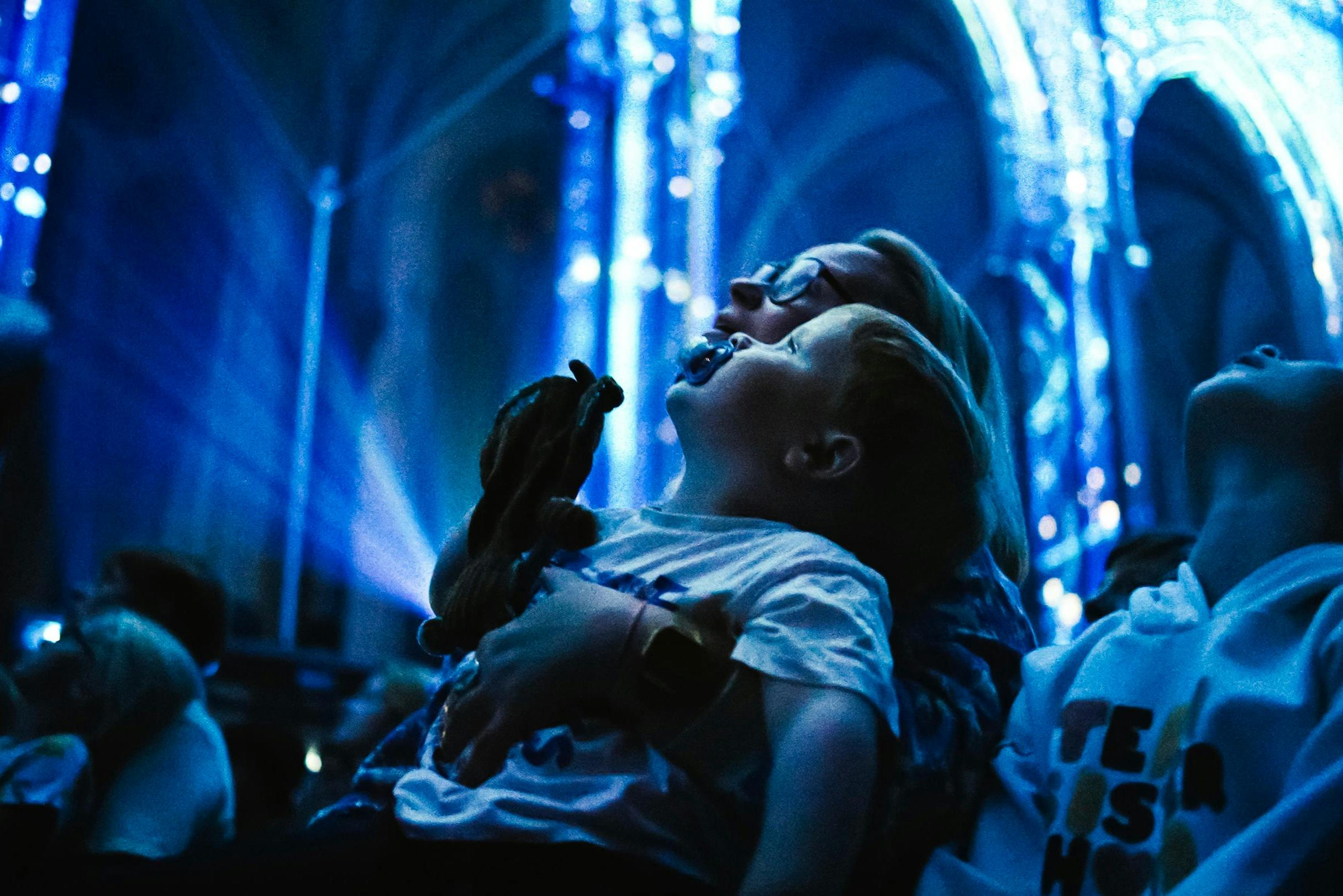An adult and child, illuminated in blue light, gaze upward inside a large, ornately lit indoor space.