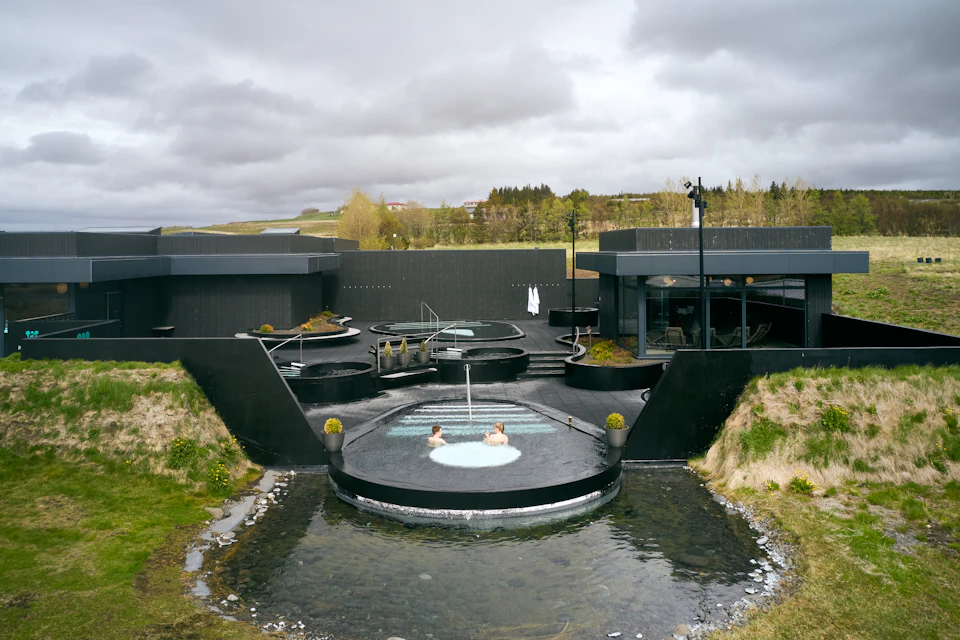 Two people relax in a circular hot tub within a modern outdoor spa surrounded by dark structures and greenery under a cloudy sky.