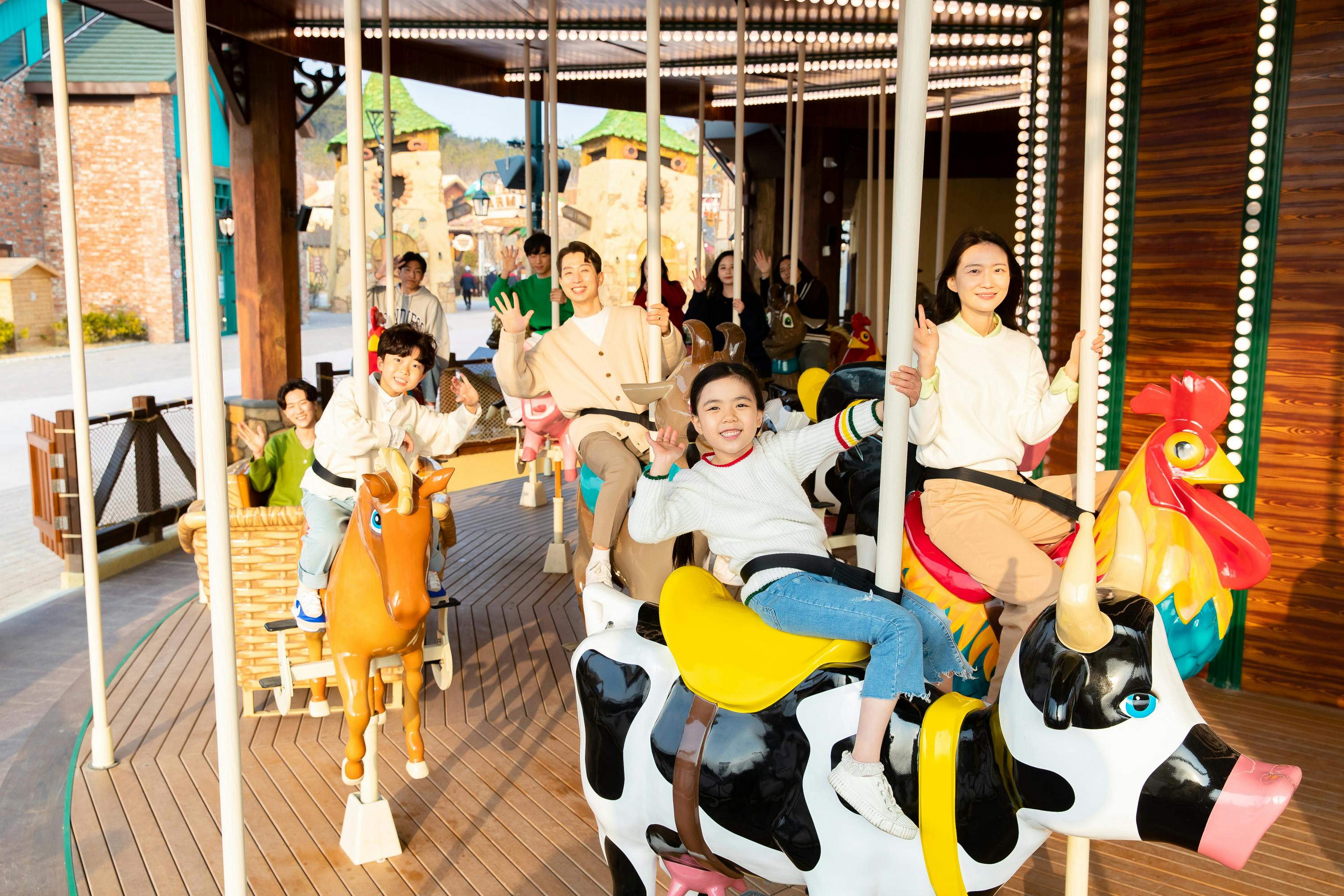Children and adults enjoying a carousel ride with animal-shaped seats in a sunny outdoor amusement park setting.