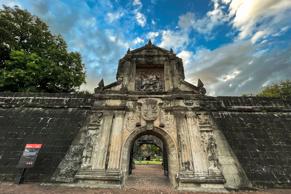 Stone gateway with intricate carvings, archway, and a relief sculpture above, surrounded by brick and greenery under a cloudy sky.