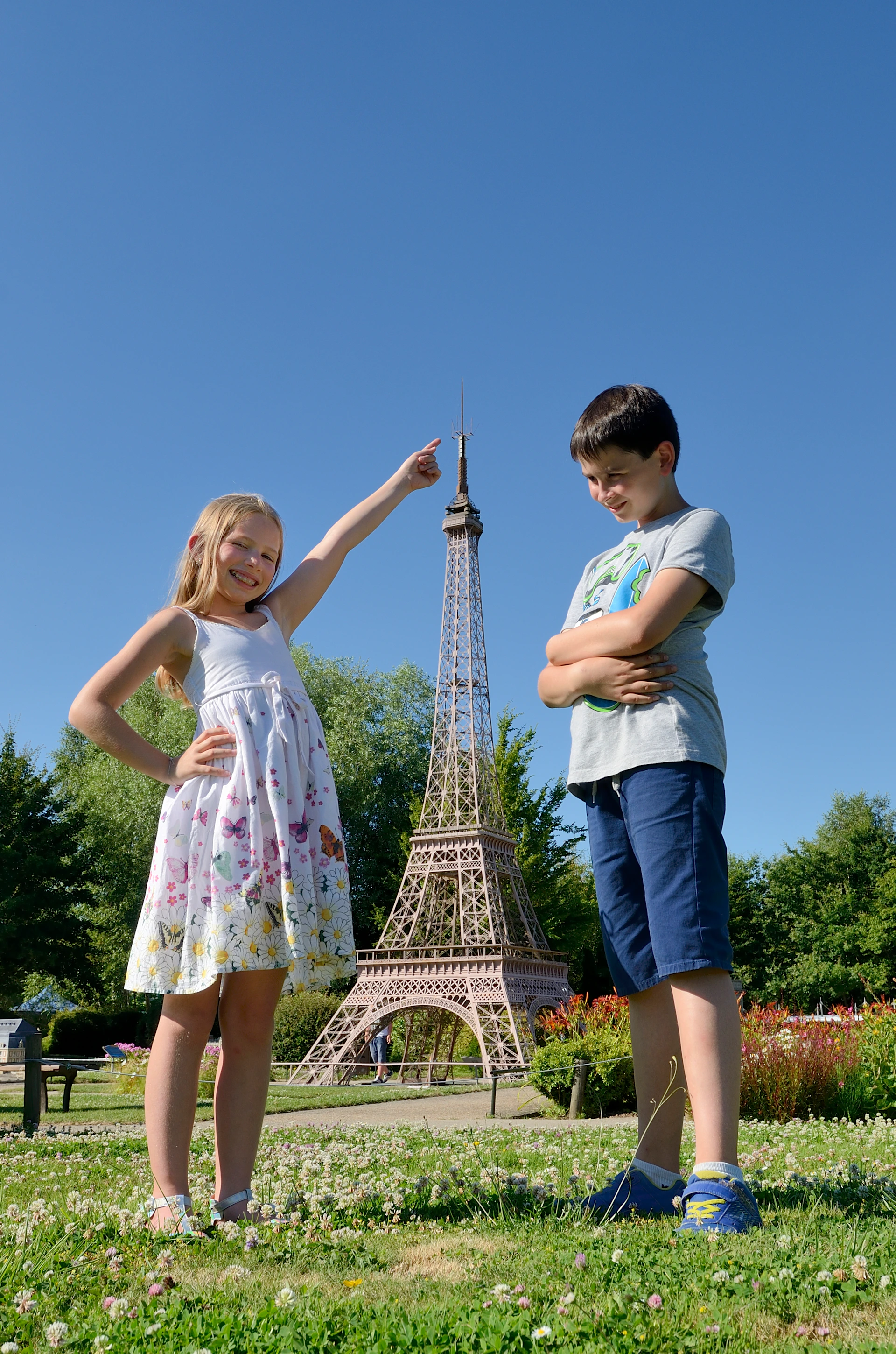 A girl points and smiles at a miniature Eiffel Tower while a boy stands beside her with arms crossed, both outdoors on a sunny day.