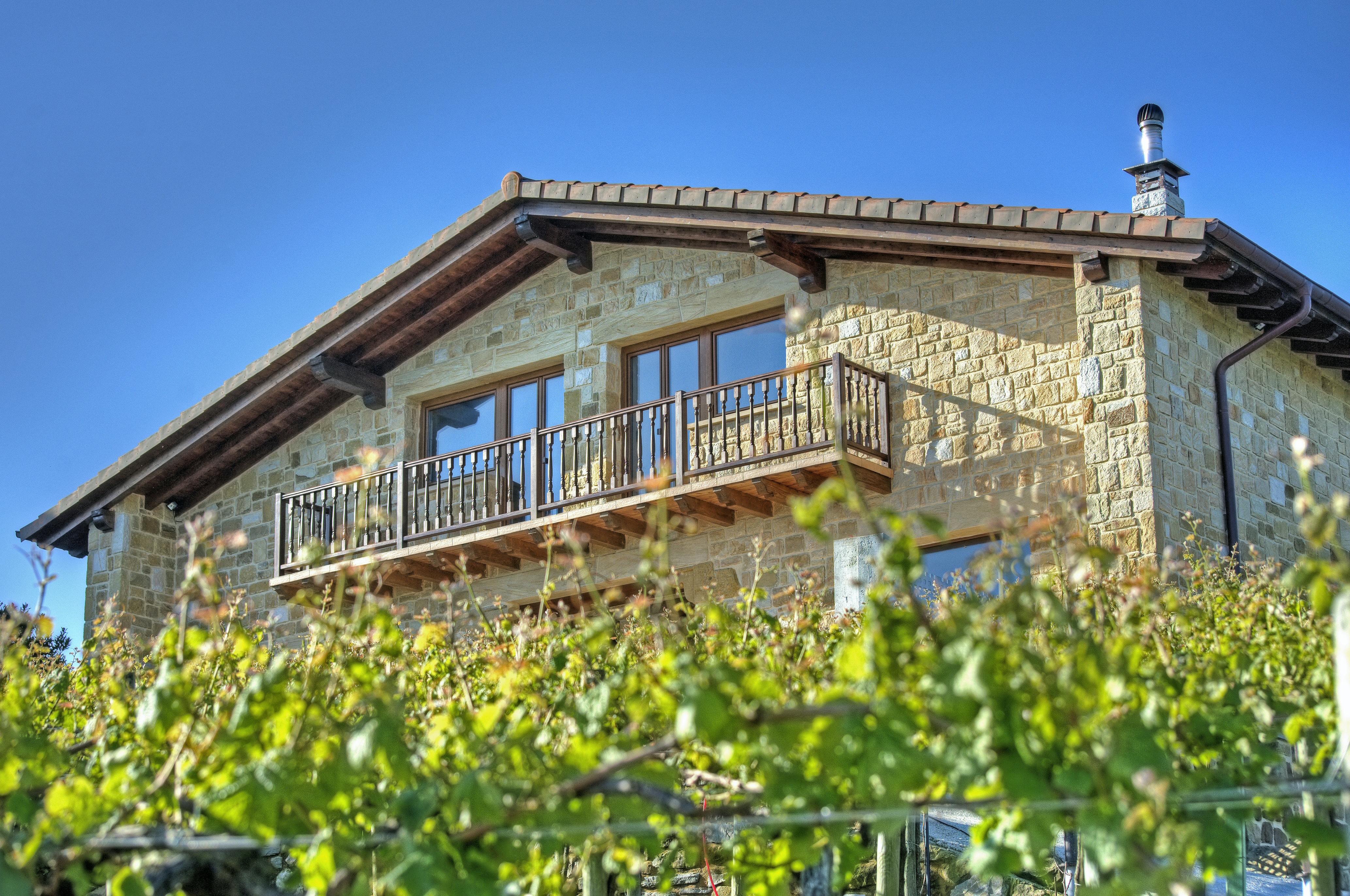 A stone house with a wooden balcony and large windows, surrounded by lush green foliage under a clear blue sky.