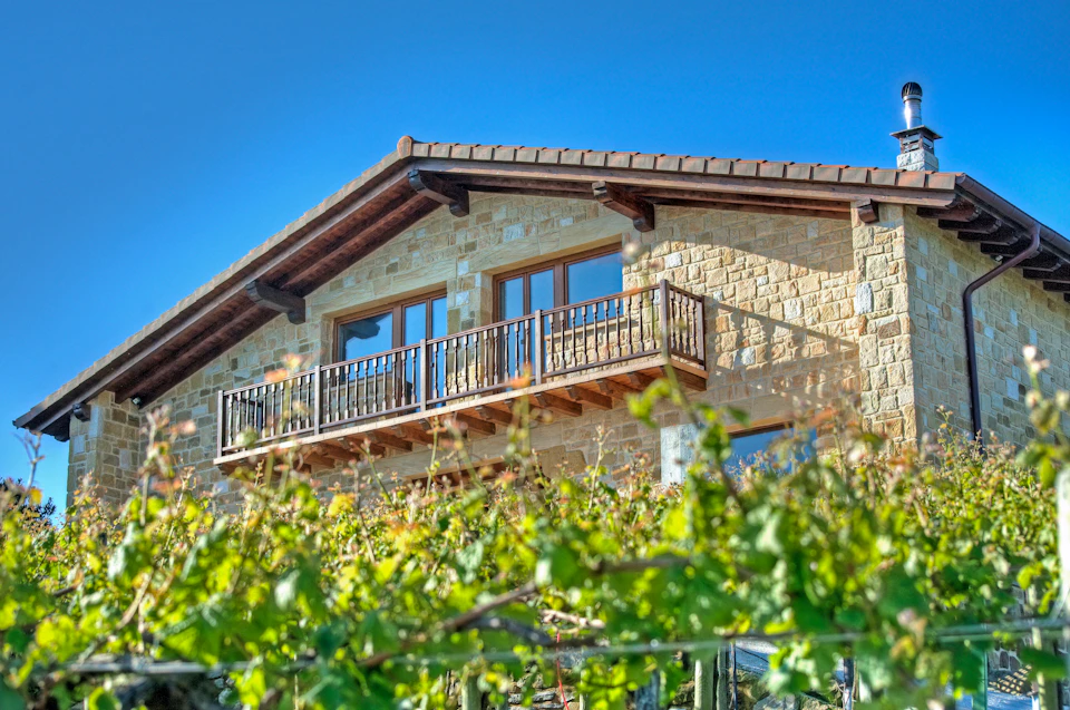 A stone house with a wooden balcony and large windows, surrounded by lush green foliage under a clear blue sky.