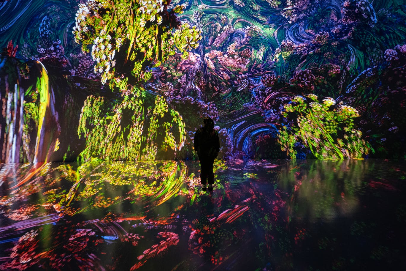 A person stands in a room with colorful, immersive digital projections of flowers and abstract patterns on walls and floor.