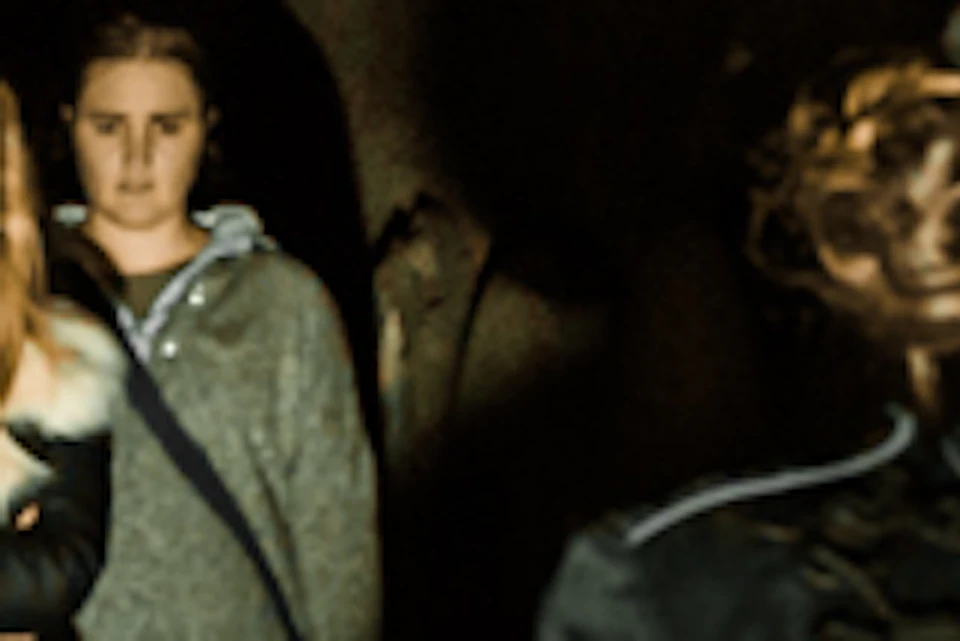 A woman in vintage attire leads a group through a dimly lit, narrow corridor lined with tombstones. The group appears intrigued.