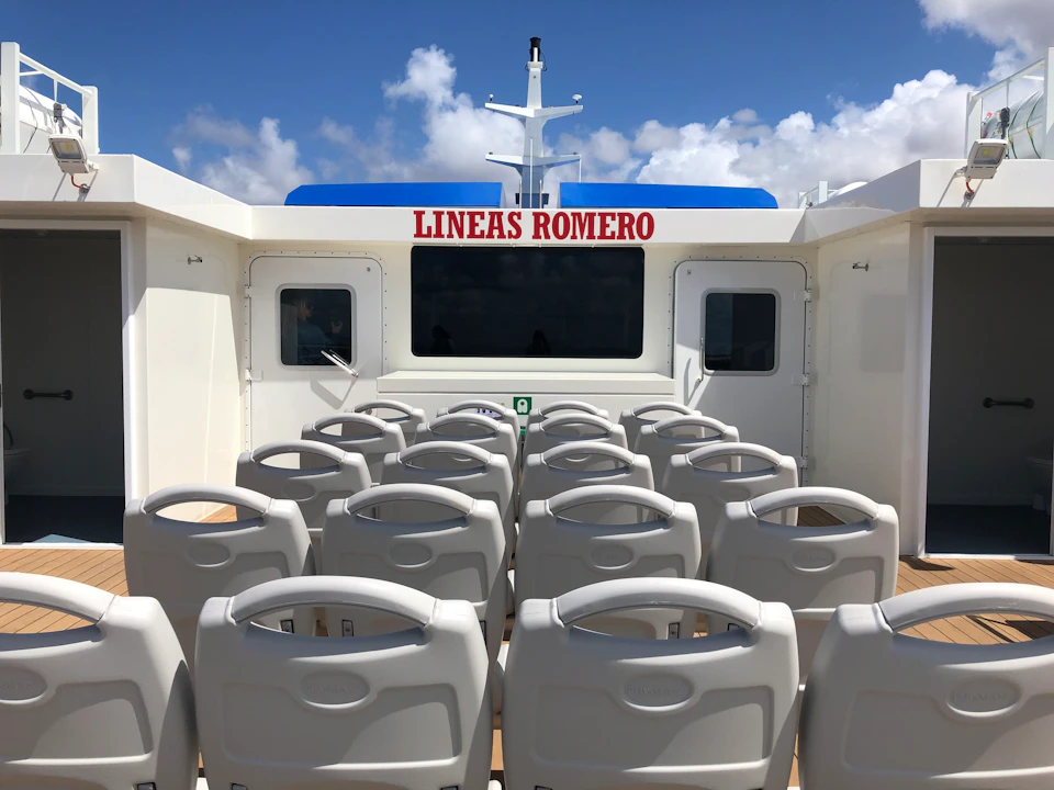 Rows of white seats on a ferry deck with "LINEAS ROMERO" signage, against a blue sky with scattered clouds.