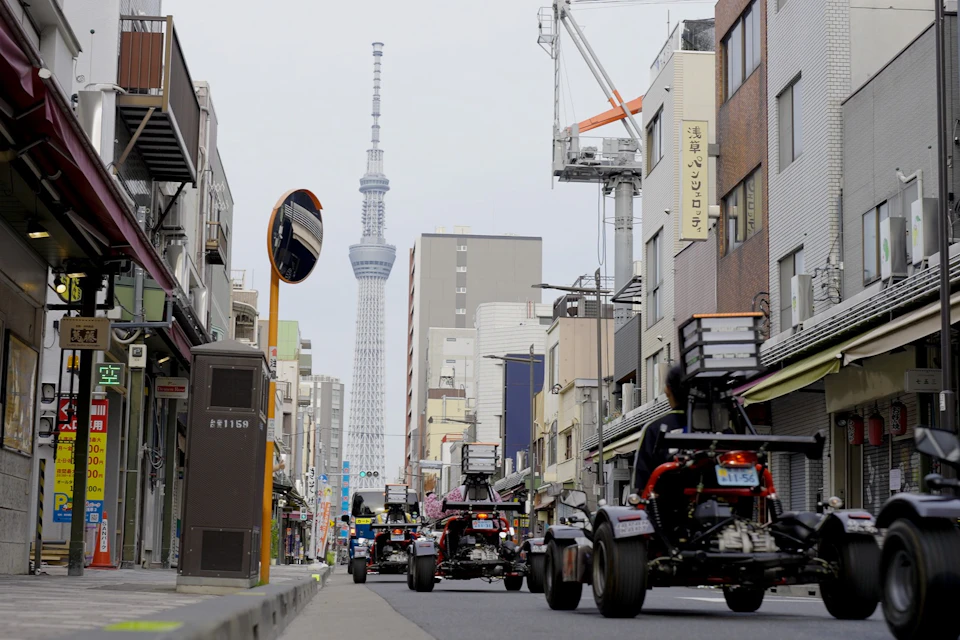 A street in Tokyo with small custom vehicles driving, buildings on both sides, and the Tokyo Skytree visible in the background.