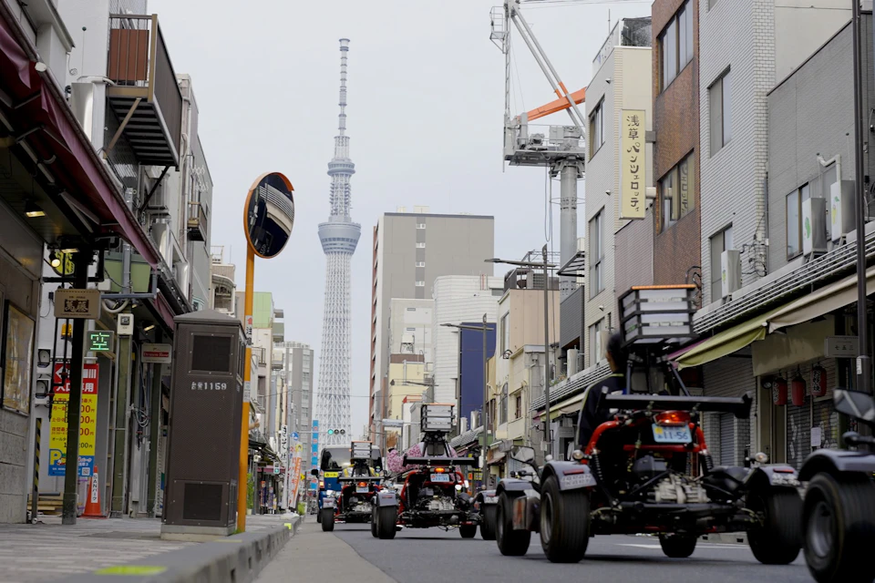 A street in Tokyo with small custom vehicles driving, buildings on both sides, and the Tokyo Skytree visible in the background.