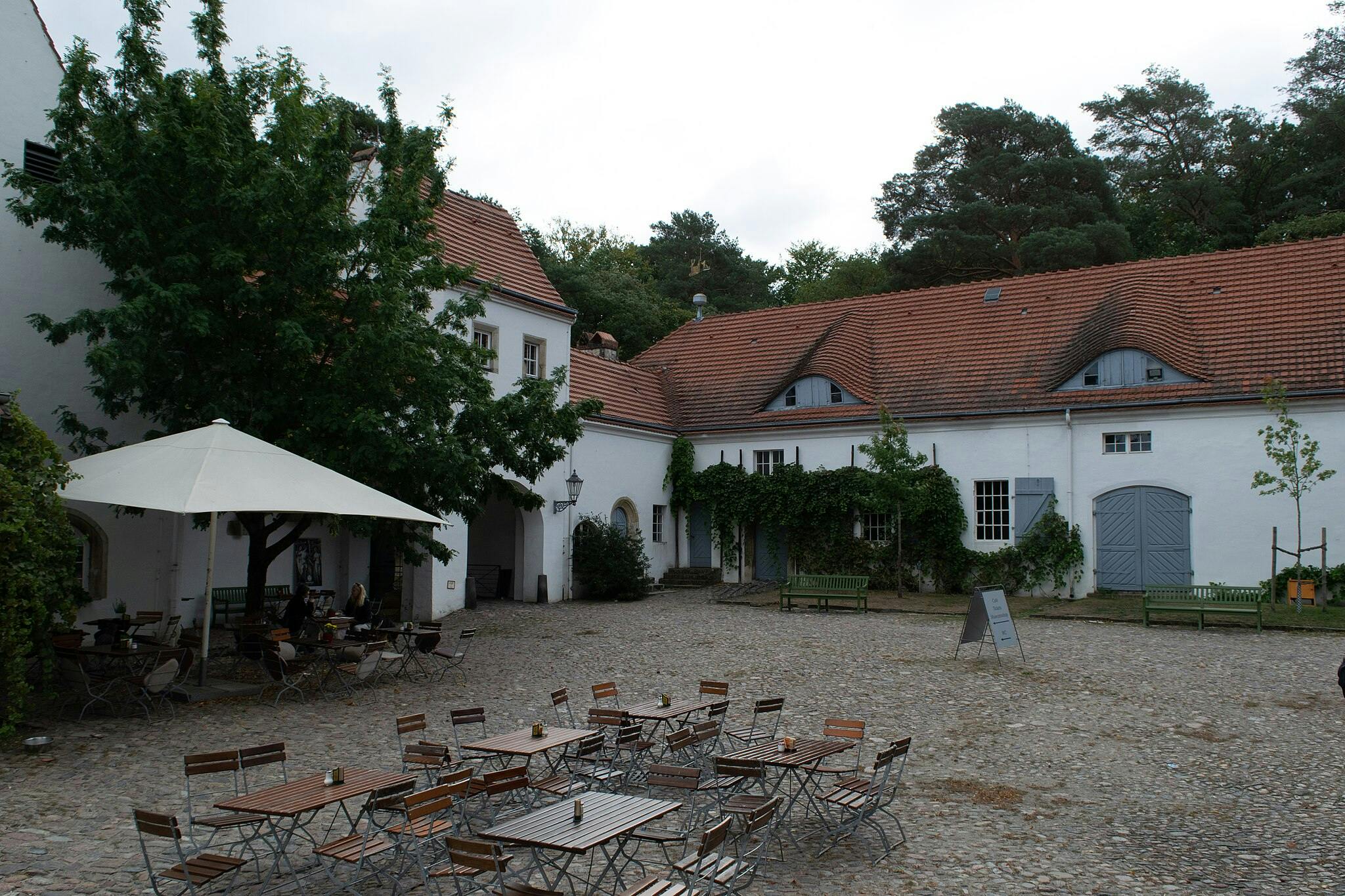 Cour extérieure avec plusieurs tables et chaises en bois, un grand parasol blanc et des bâtiments blancs entourés de toits en tuiles rouges.