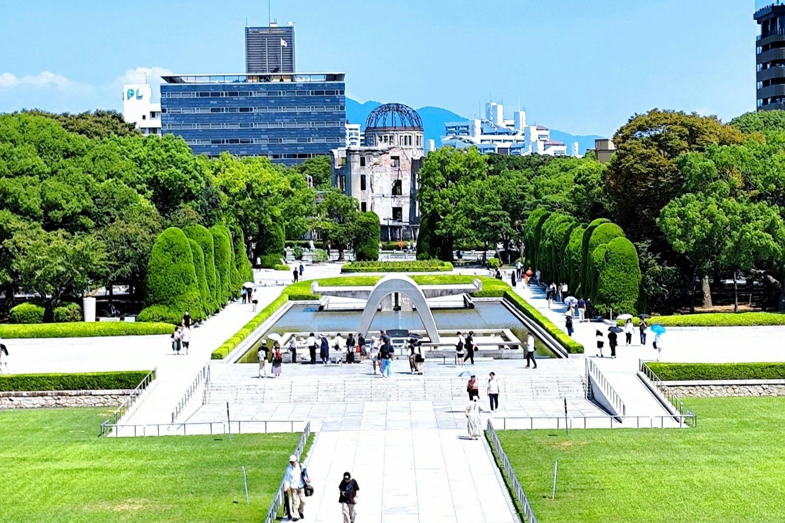 A park with a memorial arch, reflecting pool, and trees, with a domed building and cityscape in the background.