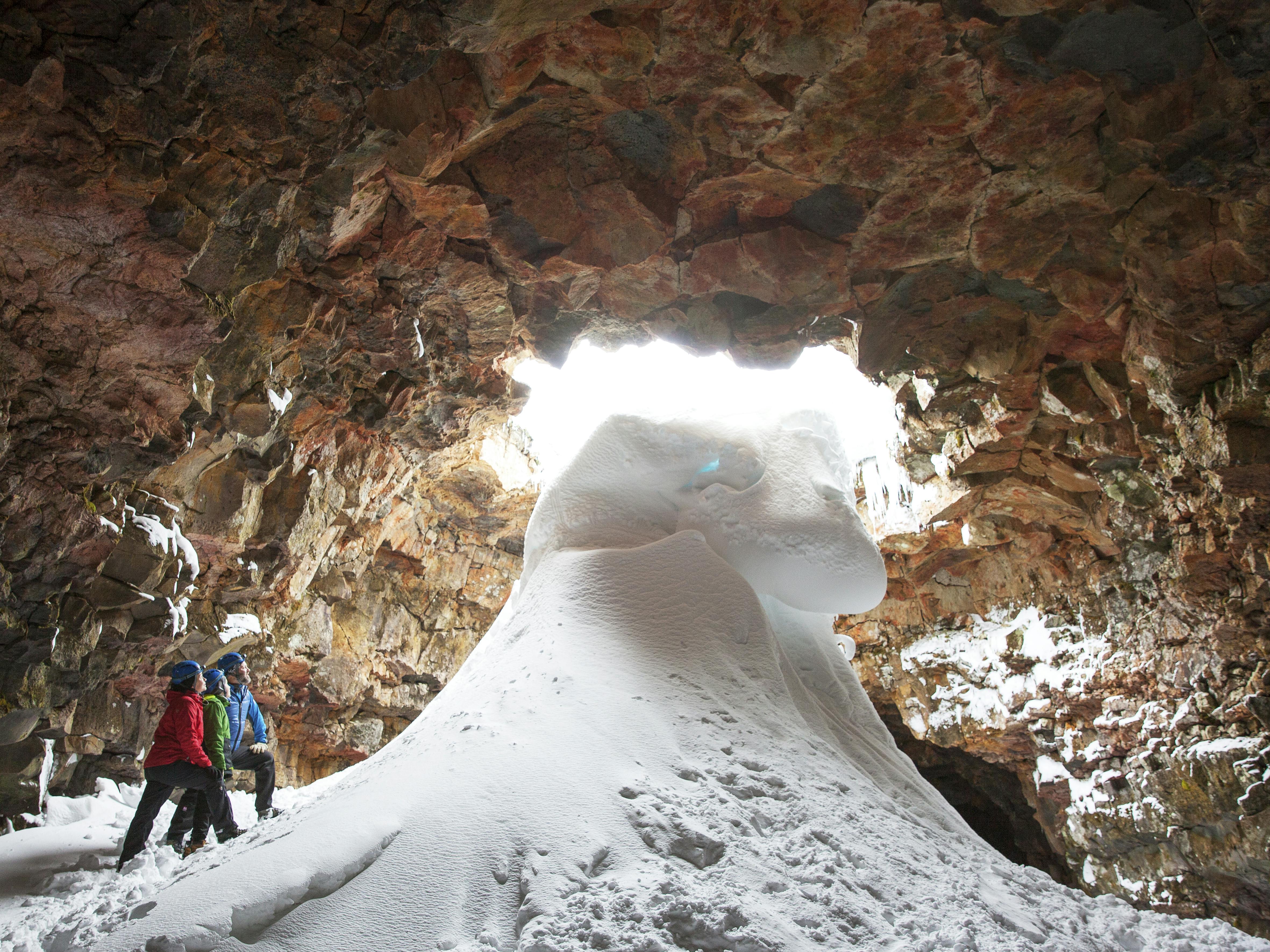 Due persone in tenuta invernale in piedi all'interno di una grotta rocciosa con un grande cumulo di neve e ghiaccio, con la luce che filtra dall'ingresso.