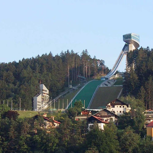 En backhoppningsramp ligger på en kulle omgiven av tät skog, med flera hus vid foten av kullen.
