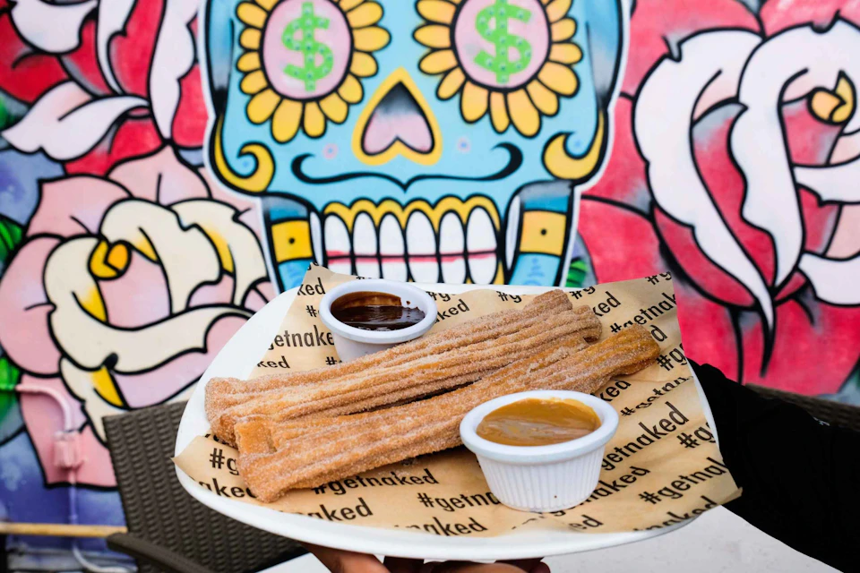 A plate of churros with dipping sauces in front of a colorful mural featuring a blue skull with money signs in its eyes.