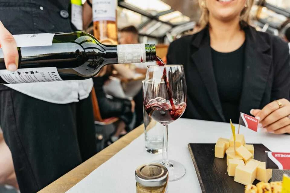 A glass of red wine being poured by a waiter for a woman, there is a platter of Dutch cheeses on the table in the foreground