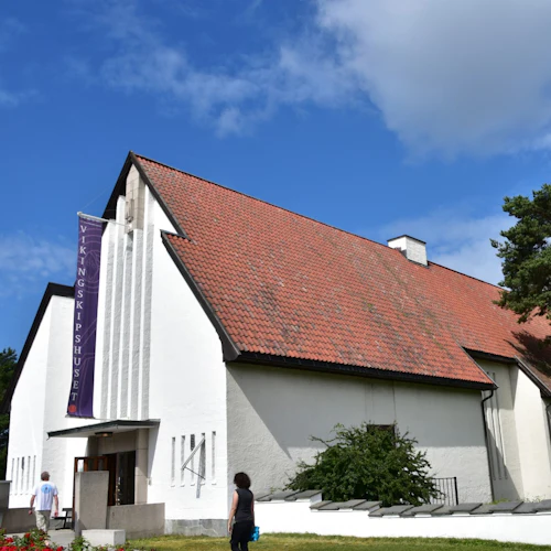 En vit byggnad med rött tegeltak och en lila banderoll med texten ”Vikingskipshuset”. Två personer går mot dess entré.