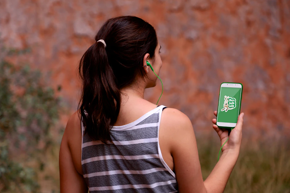 Woman with a ponytail wearing a striped tank top, holding a smartphone and wearing green earphones, with an outdoor background.