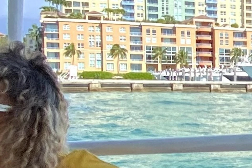 A person wearing sunglasses sitting on a boat with a cityscape of tall buildings and water in the background.