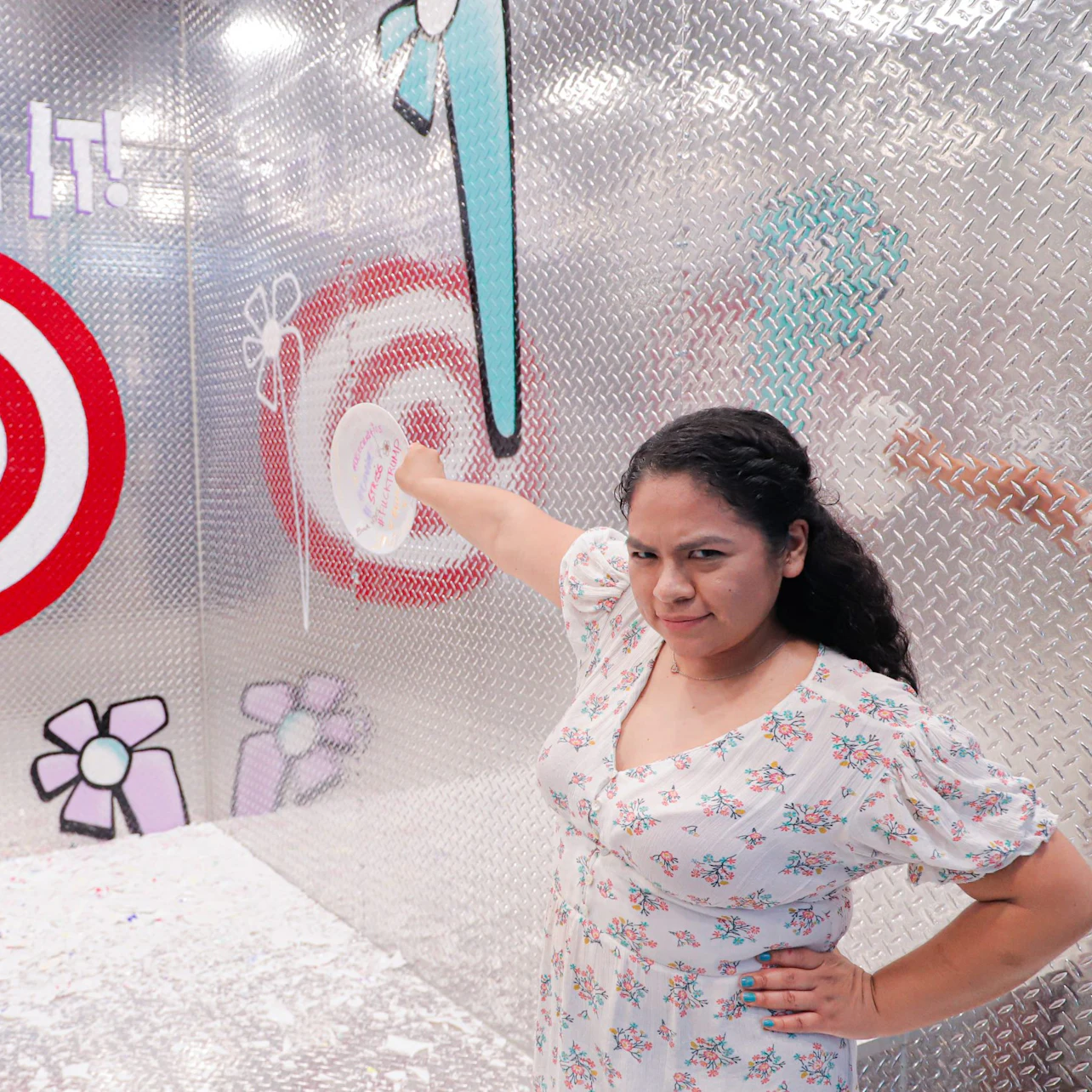 Woman in a floral dress pointing at a target on a metallic wall with cartoonish flowers and a lollipop graphic.