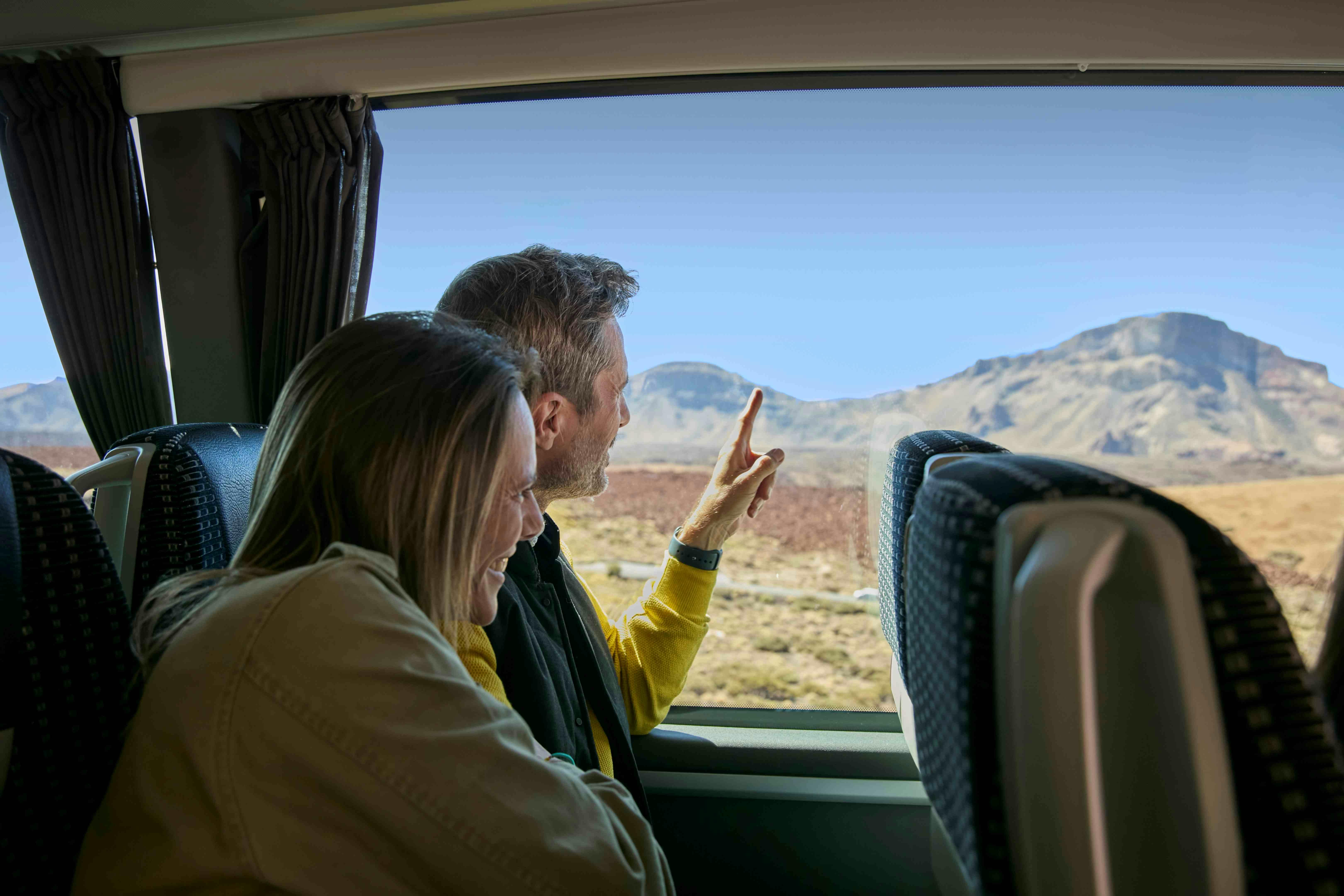 Two people sitting on a bus, looking out the window at a scenic mountain landscape. One person points at the view.