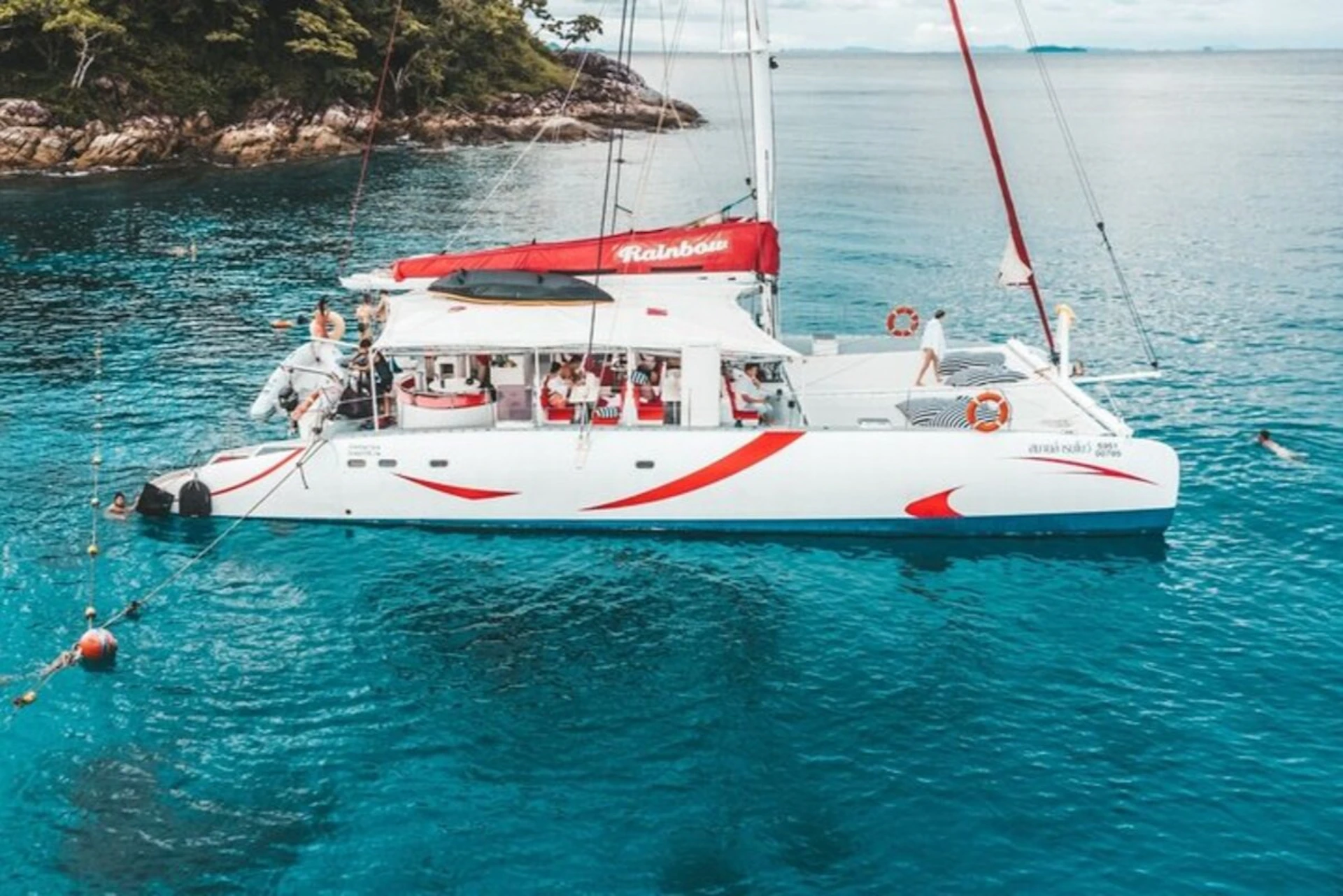 A white catamaran with red accents and passengers onboard, sailing on clear blue water near a tree-covered shoreline.