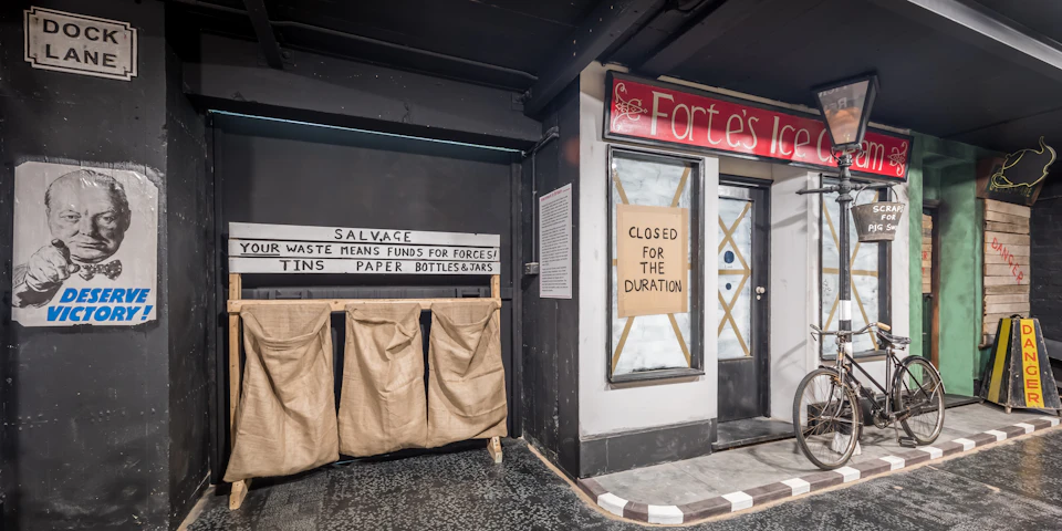 A storefront with a "Closed for the Duration" sign and a recycling area with bins for tins, paper, bottles, and jars, under a sign reading "SALVAGE."
