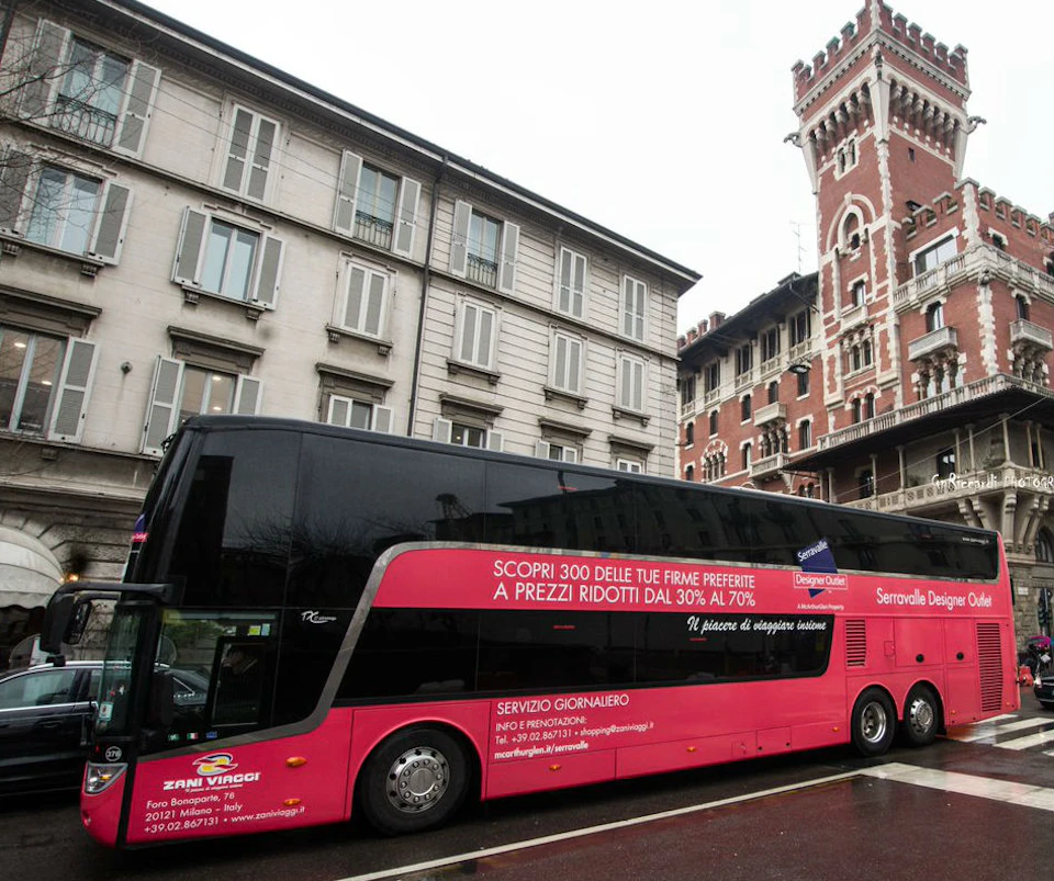 A pink double-decker bus with Italian advertisements is parked on a city street with historic buildings in the background.