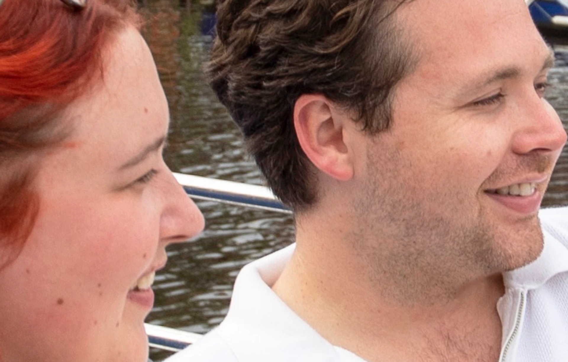 A man and a woman sit on a boat, eating food from take-out containers. A bridge, buildings, and another boat are in the background.